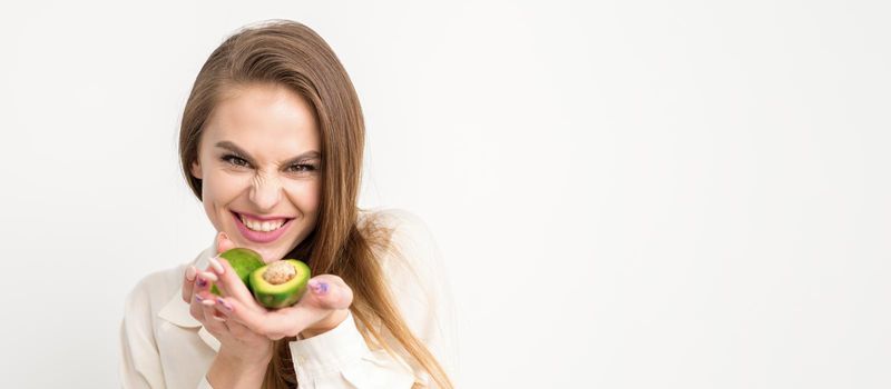 Portrait of a lovely smiling young brunette caucasian woman wearing the white shirt with long hair holding and showing avocado, standing isolated over white background