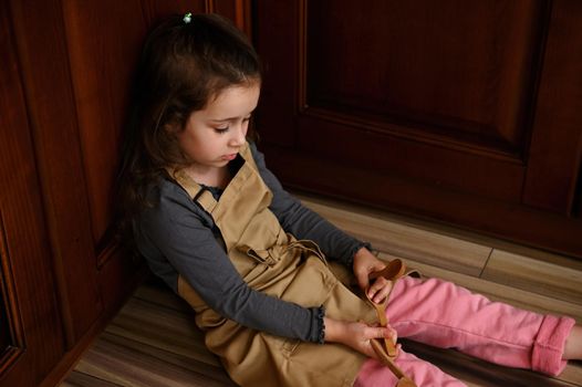 Beautiful adorable child, little Caucasian little girl in beige chef's apron playing with kitchen utensils while sitting barefoot on the floor and looking at the camera.