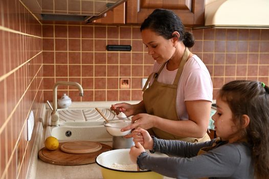 Happy family. Beautiful stunning dark-haired Hispanic woman housewife, loving mom and daughter, both in beige chef apron, preparing dough in the kitchen, enjoying cooking homemade festive pie together