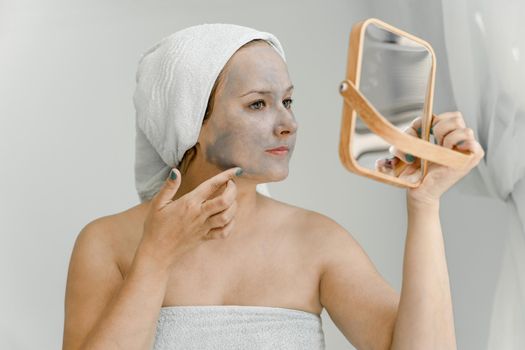 Young woman puts gray cosmetic clay mask on face while looking in mirror, her hair and body wrapped in towel. Close-up