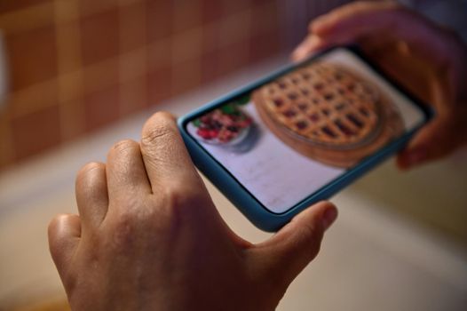 Selective focus on hands using mobile phone to photograph a homemade pie with cherries and a puff crispy crust. Smartphone in live view mode. Freshly Baked Cherry Pie with Lattice Top. Baking concept