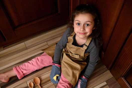 View from above to a pretty child girl, little baker confectioner in a beige chef apron and kitchen mittens, sitting on the floor and leaning against wooden kitchen cupboard, sweetly smiling to camera