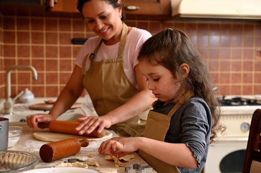 Caucasian beautiful little girl and her mom in beige aprons, playing and laughing while kneading the dough in kitchen. Homemade pastry for bread, pizza or bake cookies. Family fun and cooking concept