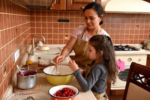 Happy multi-ethnic family, a loving mother and daughter, both wearing beige chef's aprons, baking pastries together, kneading dough for homemade festive cherry pie in the home kitchen. Family look