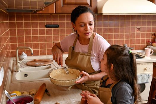 Stunning Hispanic woman, a housewife and loving mother using a fork, piercing rolled out dough in mold, teaching her cute daughter traditional family culinary while cooking a homemade pie together