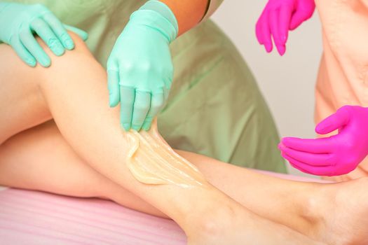 Sugaring legs. Woman legs hair removing. Hands in pink and blue rubber gloves of two beauticians apply sugar paste on female feet