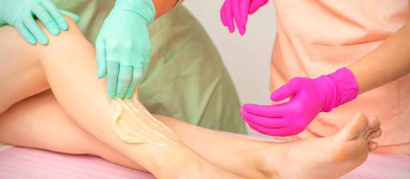 Sugaring legs. Woman legs hair removing. Hands in pink and blue rubber gloves of two beauticians apply sugar paste on female feet