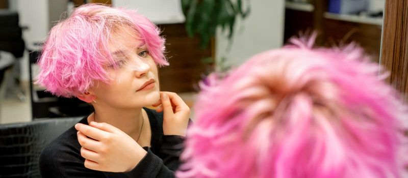 The beautiful young caucasian woman with a new short pink hairstyle looking at her reflection in the mirror checking hairstyle in a hairdresser salon