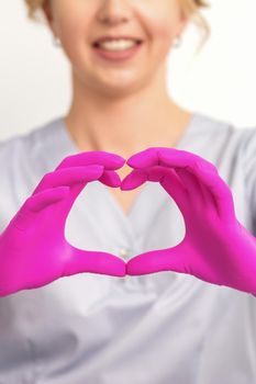 A smiling caucasian woman doctor wearing pink gloves in uniform showing the symbol of a heart against a white background