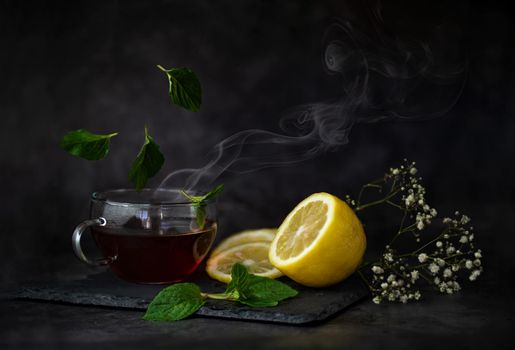 winter still life with a cut bright yellow lemon, a cup of fragrant tea and mint leaves floating around them on a table on a dark background High quality photo