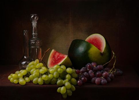 still life with a cut watermelon with different varieties of green and black grapes and a couple of decorative bottles on the table on a dark background High quality photo
