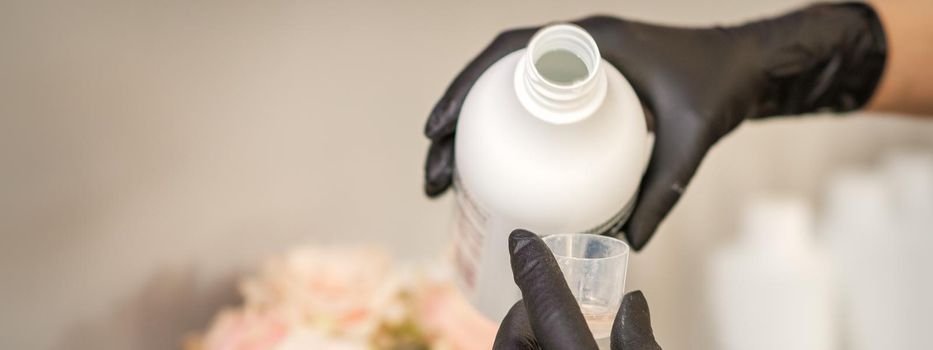 A hairdresser in black gloves is preparing hair dye with a bottle in a hair salon, close up