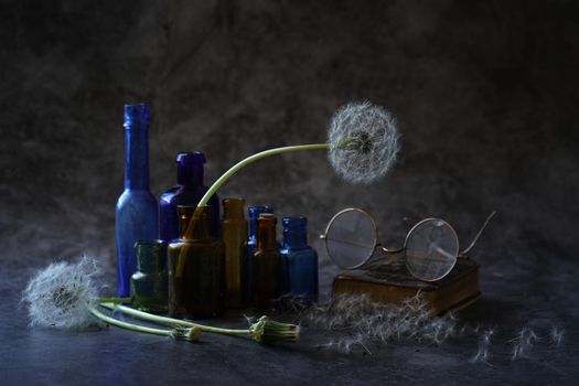 a still life consisting of dandelions with white fluffy heads multicolored small bottles and antique glasses of the late 19th and early 20th century on a table on a dark background. High quality photo