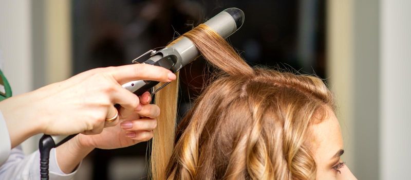 Hairdresser makes curls with a curling iron for the young woman with long brown hair in a beauty salon