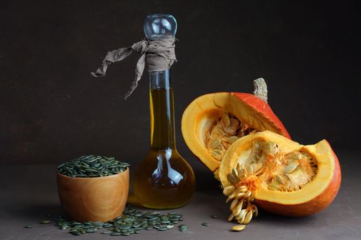 still life consisting of sunflower oil in a decorative bottle of two halves of pumpkin and lying around dried shelled fried dark pumpkin seeds on a table on a dark background. High quality photo