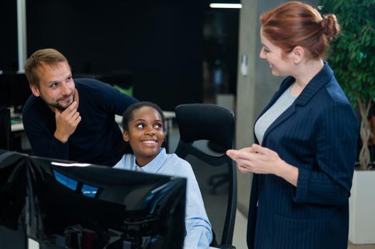 Colleagues look at the monitor and decide working moments. African young woman, caucasian man and red-haired caucasian woman communicate in the office