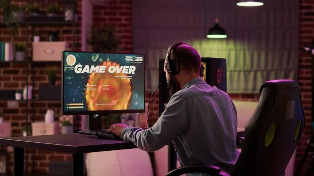 Gamer feeling disappointed after losing difficult level in online space shooter on gaming pc in home living room. Man playing internet multiplayer game sad and bored after failing tournament.