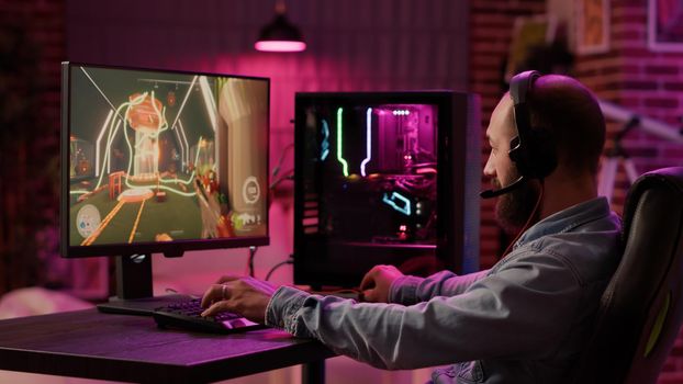 Static tripod shot of man using pc setup playing multiplayer online action game talking to team on headset. Gamer sitting in gaming chair streaming first person shooter while explaining gameplay.
