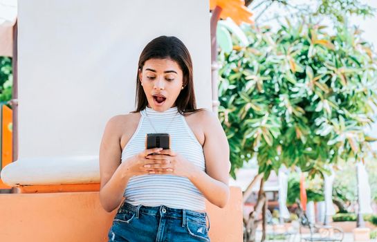 Surprised person looking at cell phone outdoor. Girl with cell phone with a surprised face leaning on a wall. Young woman with the cell phone with a gesture of surprise with open mouth outside