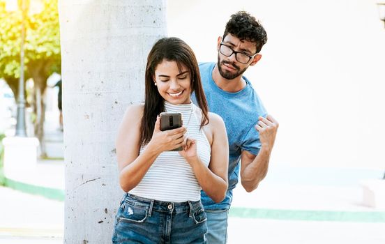 Suspicious man spying on his girlfriend with his cell phone, Jealous boyfriend spying on his girlfriend's cell phone in the park. Jealous man spying on his girlfriend texting on the cell phone