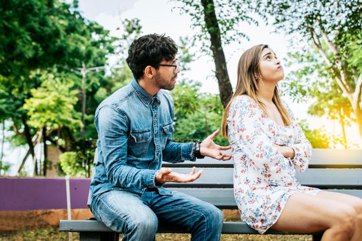 Upset couple arguing in a park. A young couple arguing sitting on a park bench, Concept of couple problems and crisis. Man arguing with his girlfriend sitting in a park