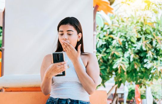 Girl with cell phone with a surprised face leaning on a wall. Young woman with the cell phone with a gesture of surprise covering her mouth outdoors. Surprised person looking at cell phone