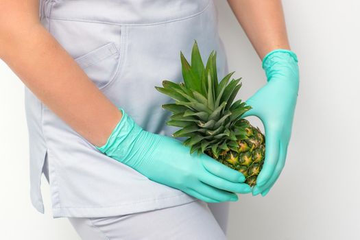 Young beautician wearing blue gloves in uniform with pineapple covers an intimate area on a white background, bikini zone depilation concept