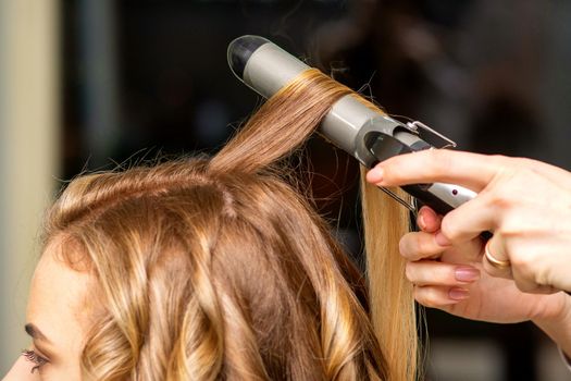 Hairdresser makes curls with a curling iron for the young woman with long brown hair in a beauty salon