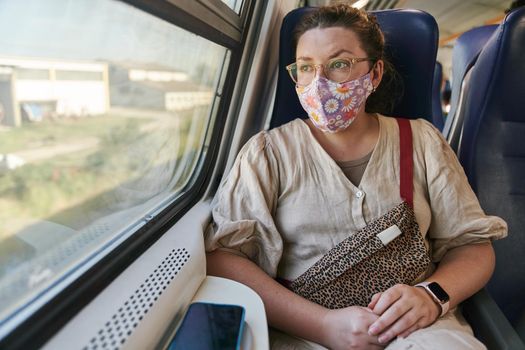 A girl in glasses and a medical mask riding a train and looking out the window. High quality photo