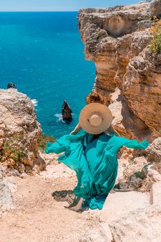 A girl with loose hair in a long mint dress descends the stairs between the yellow rocks overlooking the sea. A rock can be seen in the sea. Sunny path on the sea from the rising sun.