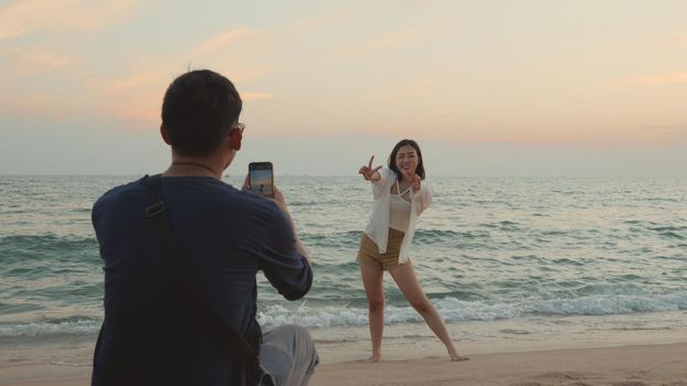 Happy man using smartphone camera making photo to woman at the beach and having fun together on beach in holiday vacation time at sunset, Young Asian woman and man in summer travel outdoor