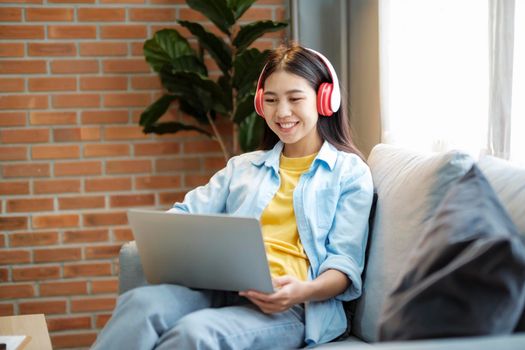 Young asian woman studying, online learing, and listening to music with headset while sitting on couch at home. Listening to podcast, audio book, smiling, and, watching videos using laptop.