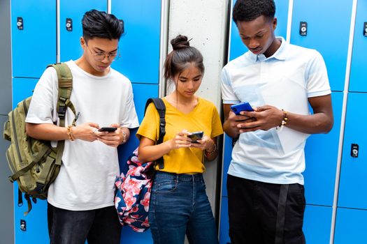 Group of multiracial teen classmates using mobile phone with disinterest on each other in high school corridor. Technology addiction in alienated lifestyle concept.