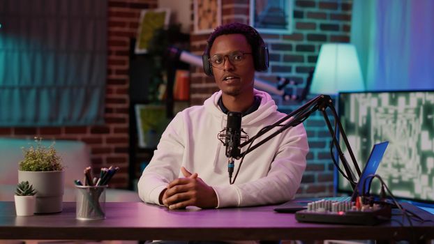 African american online radio host reading fan mail from laptop talking using microphone in home studio. Social media content creator answering questions from listeners while recording podcast.