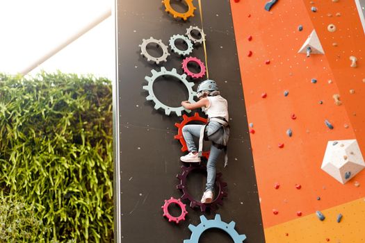 Child climbing on wall in amusement centre. Climbing training for children. Little girl in dressed climbing gear climb high. Extreme active leisure for kids. Caucasian little girl 5 years old.