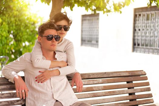 Smiling beautiful woman and her handsome boyfriend. Woman embrace summer day. Happy cheerful couple in sunglasses walking old city park. Couple posing on the street near green tree