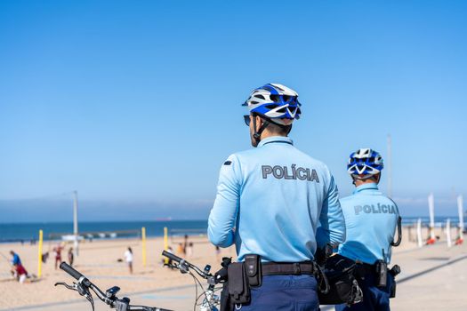 Two policeman patrolling seaside promenade on bicycles. People are sunburning on the city public beach on the Atlantic shore. Bike police patrol on the ocean beach