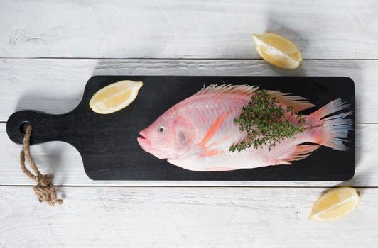 raw fresh pink tilapia fish lies on a board sprinkled with small red and green hot peppers, pink Himalayan salt and garnished with fresh curry sprigs, on white wooden planks. High quality photo