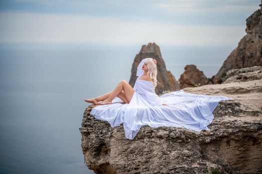 The blonde sits on a rock above the sea. With long hair on a sunny seashore in a white dress, rear view, silk fabric flutters in the wind. Against the backdrop of blue skies and mountains