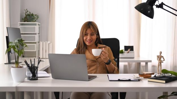 Creative female manager drinking coffee and reading email on her laptop computer.