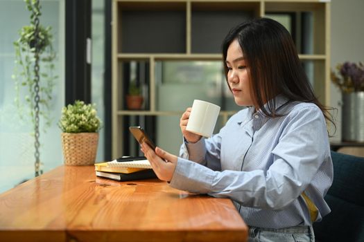 Asian woman employee having coffee break in corporate office and using mobile phone.