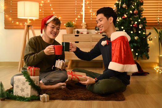 Two happy man next to a decorated Christmas tree and enjoying drinking hot chocolate together.