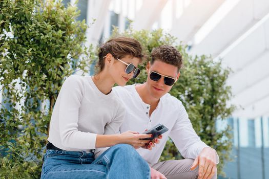 Young modern couple use smartphone sitting bench outdoor in office district. Beautiful woman and handsome man in sunglasses holding smartphone in hand looking device screen and smile.