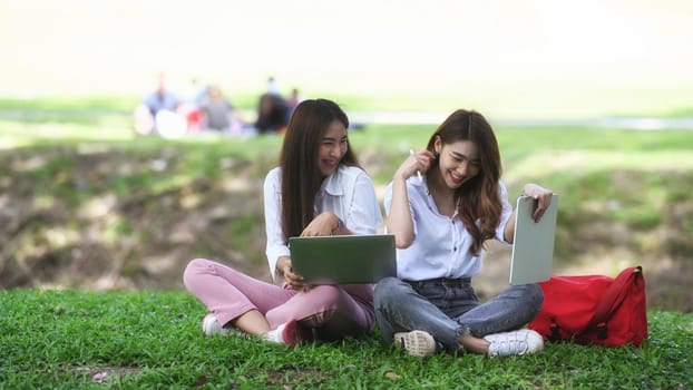 Two cheerful young women sitting on grass in the park and working with computer laptop.