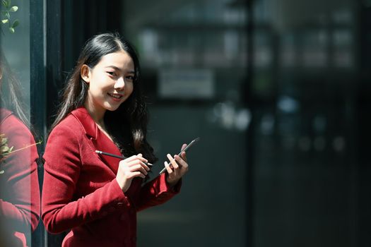 Attractive businesswoman standing in modern office and using smart phone.