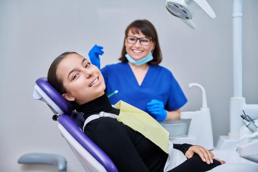 Portrait of young female patient with toothy smile looking at camera sitting in dental chair with dentist doctor. Dentistry, hygiene, treatment, dental health care concept
