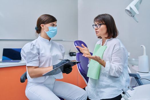 Dentist and woman patient in dental clinic, doctor nurse writing on clipboard consulting, middle aged female talking about disturbing dental problem. Dentistry, treatment, dental health care concept