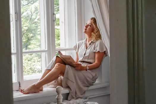 A middle-aged woman in a cream dress sits mysteriously and looks out the window on the windowsill. Green trees outside