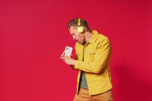 Joyful man sing while recording his voice using smartphone. Singing handsome man enjoying his favorite song using phone and wireless headphones wearing jeans yellow jacket isolated on red background.