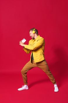 Full portrait of singing handsome man enjoying his favorite song using phone and wireless headphones wearing casual jacket isolated on red background. Man sing while recording his voice.